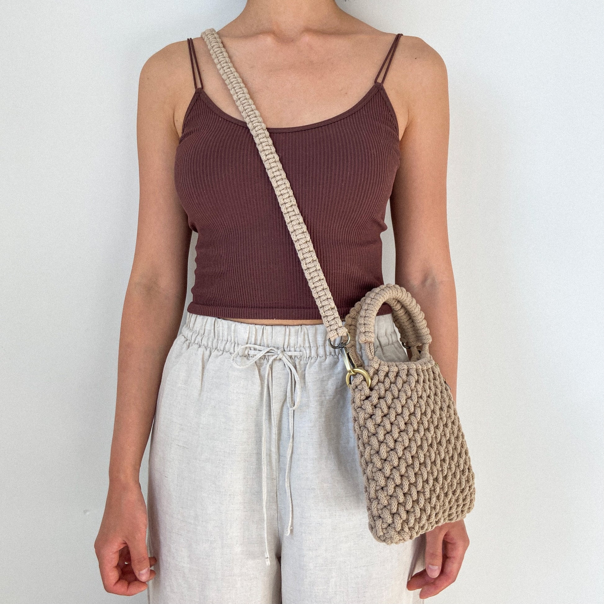 Person wearing a brown ribbed tank top and white pants, holding a beige woven handbag against a plain background.