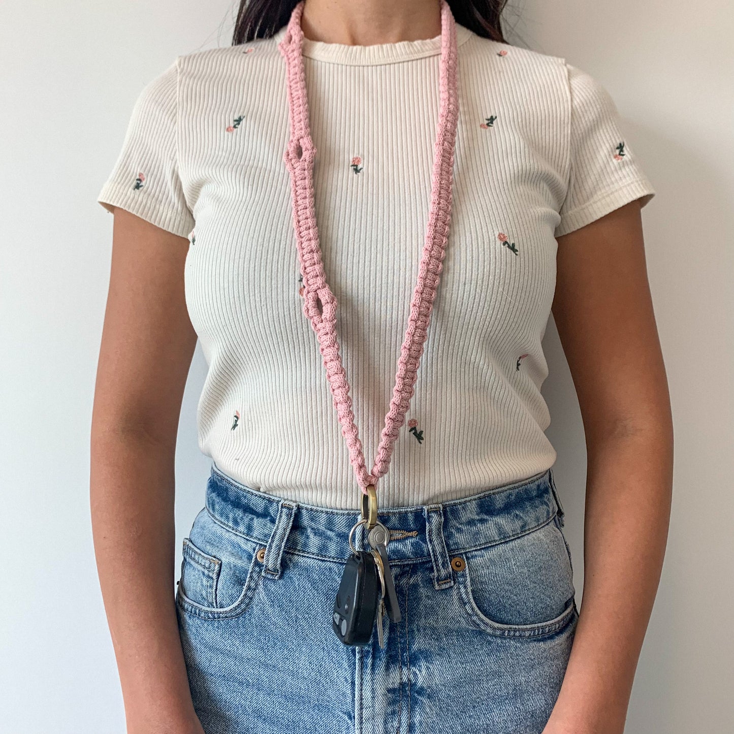 Person wearing a white t-shirt with embroidered designs and blue jeans, holding a macrame lanyard.