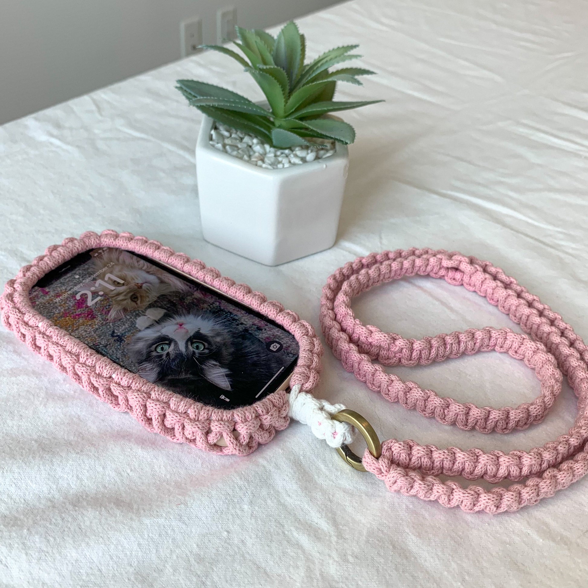 Pink crocheted phone case with a lanyard on a white surface with a small plant in the background.